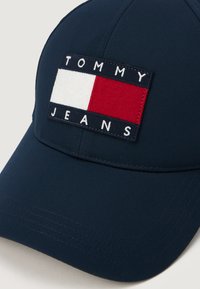 Marineblå baseball cap laget av stoff, med en rektangulær lapp som har teksten "TOMMY JEANS" i røde, hvite og blå farger.