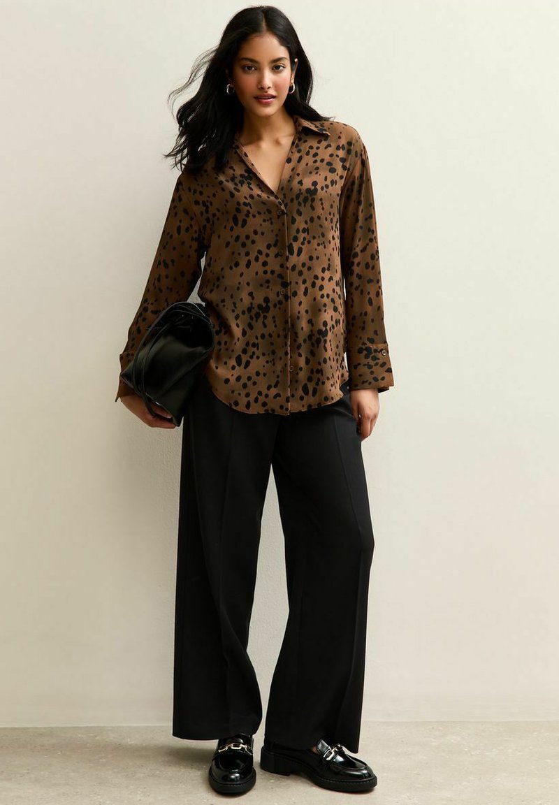 Camisa marrón de manga larga con botones y manchas de leopardo negras, combinada con pantalones de pierna ancha negros y mocasines negros brillantes. Bolso de mano negro.