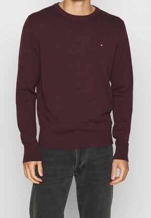 Homme portant un pull bordeaux ajusté à manches longues avec un petit logo sur la poitrine et un jean gris foncé, debout devant un fond uni.