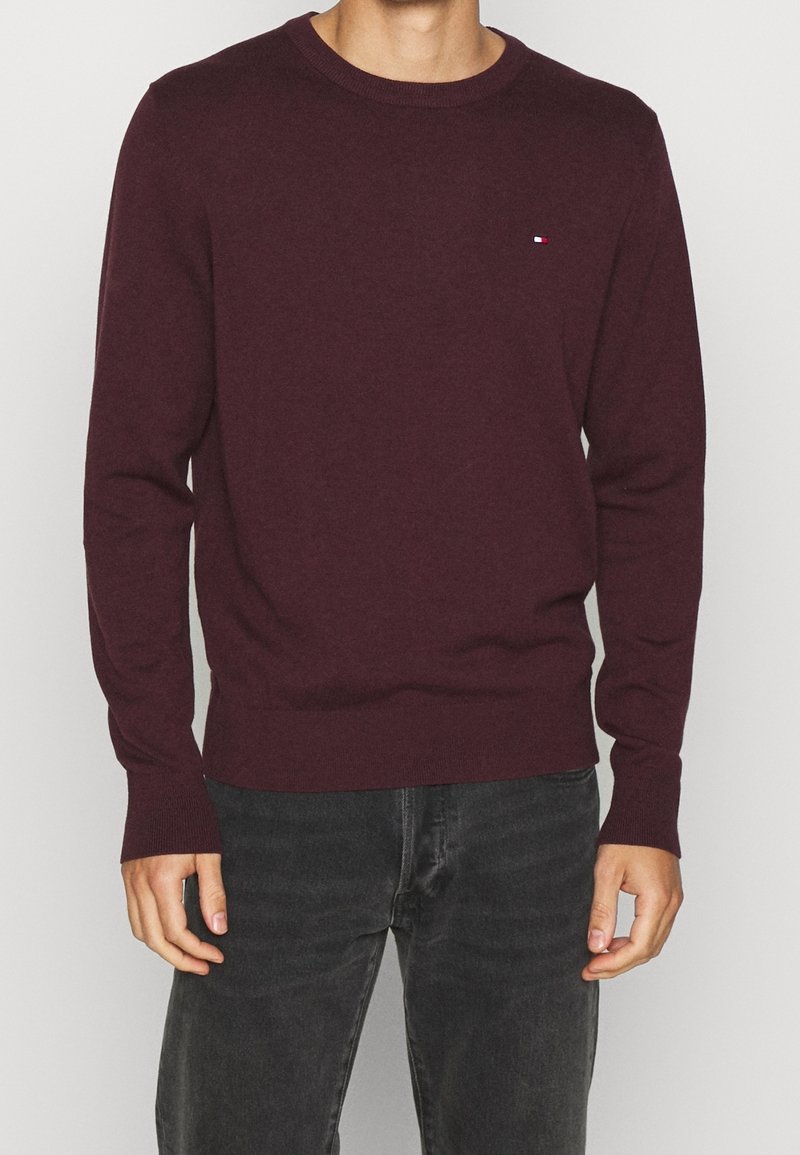 Homme portant un pull bordeaux ajusté à manches longues avec un petit logo sur la poitrine et un jean gris foncé, debout devant un fond uni.
