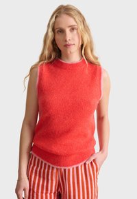 Röd ärmlös stickad tröja med rund halsringning och mjukt material, med en kontrasterande rosa nederkant. Matchad med randiga byxor.