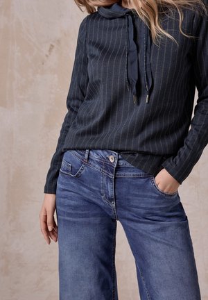 Frau trägt eine dunkle, gestreifte Bluse mit Kordelzügen und blaue Jeans, mit einer Hand in der Tasche vor neutralem Hintergrund.