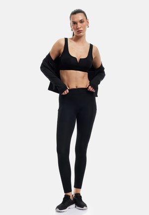 Vrouw met een zwarte sportbeha, zwarte legging, zwart jasje dat van de schouders glijdt, en zwarte sportschoenen, staand tegen een effen achtergrond.