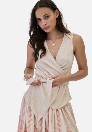 Joven con cabello largo y oscuro que lleva un top cruzado sin mangas de color beige y una falda estampada, atándose la cinta de la cintura del top.