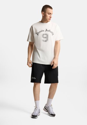 Młody mężczyzna w białej koszulce "Prosto Infinity 9", czarnych szortach "Prosto", białych skarpetkach i czarno-białych trampkach, stoi i patrzy w dół.