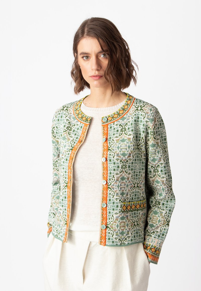 Tkana zelena jakna s složenim uzorcima, narančastim detaljima i okruglim dugmićima. Neuredan kroj, prikazana preko lagane majice.