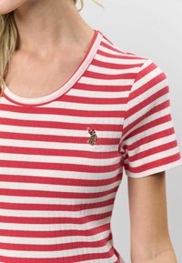 Sarkana un balta svītrota īsā piedurkņu krekls ar ribotu tekstūru un apaļu izgriezumu. Ievērojama maza izšūta detaļa uz krūtīm.