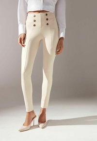 Pantalons beiges taille haute avec une texture lisse, cinq accents de boutons dorés et une coupe ajustée. Assortis à des escarpins nude à bout pointu.