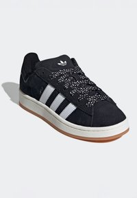 Crne suède tenisice s tri bijele pruge i crno-bijelim uzorcima na vezicama. Imaju gumeni potplat i teksturirane detalje na gornjem dijelu.