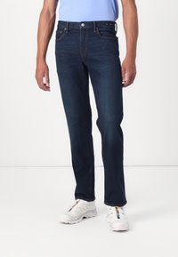 Dark blue denim jeans with a straight-leg fit, front pockets, and contrasting stitching, paired with white athletic shoes.
