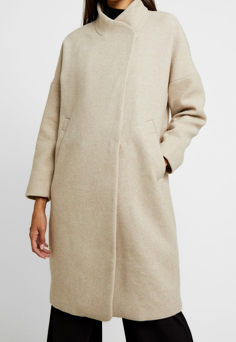 Femme portant un manteau beige mi-longueur jusqu'aux genoux avec un col haut et des poches latérales, associé à un pantalon noir et un top noir en dessous.