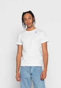 Camiseta de algodón blanca con cuello redondo y mangas cortas; presenta un pequeño logo azul y rojo en el lado izquierdo del pecho; combinada con jeans de color azul claro.