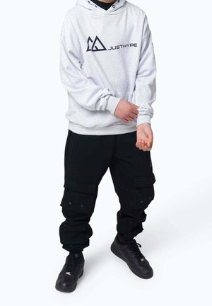 Person, der justerer ærmet på en lysegrå hættetrøje med "JUSTHYPE"-logo, iført sorte cargobukser og sorte sneakers, stående mod en ensfarvet baggrund.