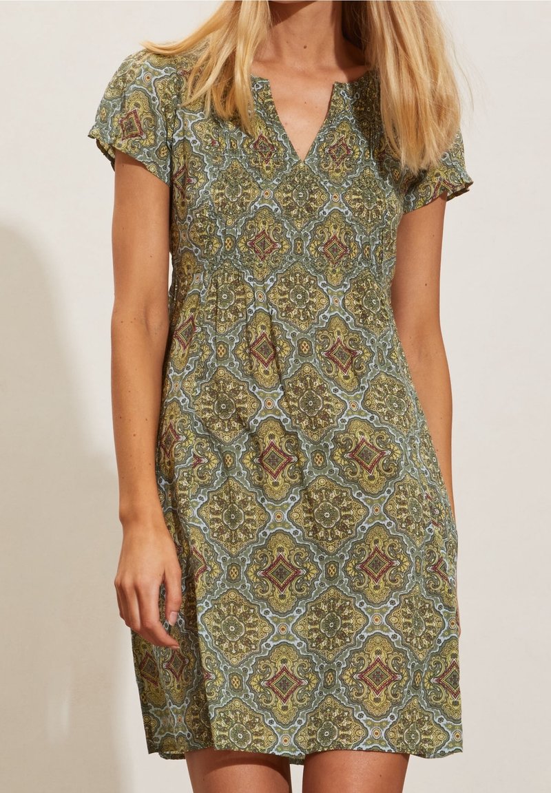 Femme portant une robe à manches courtes, longueur genou, avec un motif géométrique orné vert, jaune et rouge et un décolleté en V sur fond uni.
