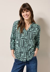 Mintgrünes Hemd mit geometrischem schwarzen Muster, Knopfleiste, lässiger Schnitt und langen Ärmeln, kombiniert mit blauen Jeans.