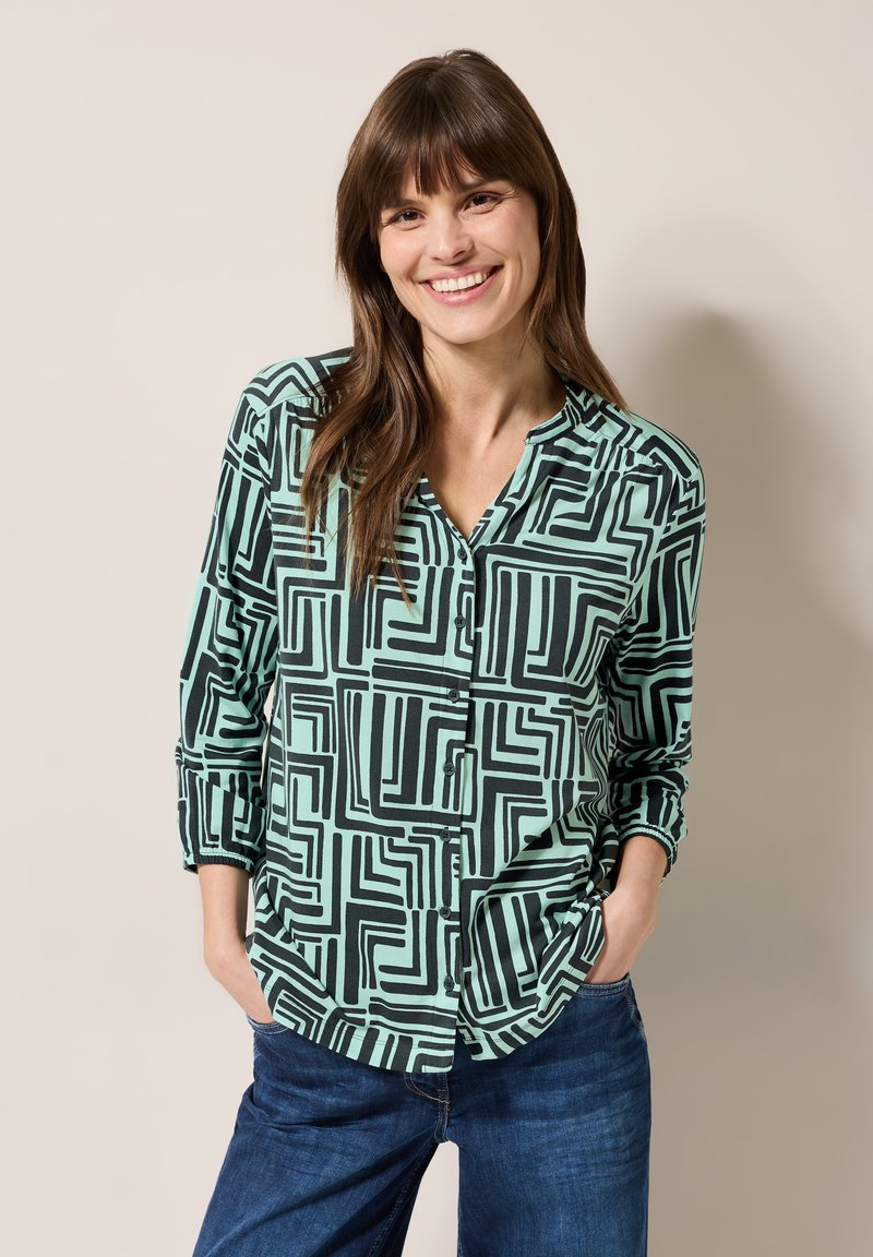 Mintgrünes Hemd mit geometrischem schwarzen Muster, Knopfleiste, lässiger Schnitt und langen Ärmeln, kombiniert mit blauen Jeans.