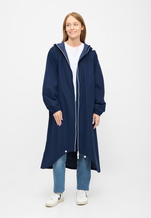 Ung kvinde smilende iført en lang marineblå hætte-regnjakke, hvid skjorte, blå jeans og hvide sneakers, stående på en ensfarvet baggrund.