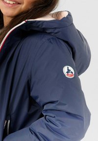 Giubbotto imbottito blu navy con cappuccio, dotato di un'elegante fodera rosa chiaro e di un logo circolare sulla manica. Chiusura frontale con zip.