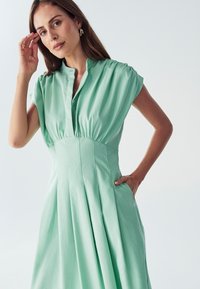 Mäta zelené šaty s krátkymi rukávmi, golierovým výstrihom, plisovanou sukňou a bočnými vreckami. Jemný materiál s hladkou textúrou a priliehavým pásom.