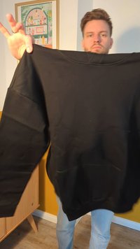 Black sweatshirt with long sleeves, ribbed cuffs, and a front pocket. Material appears soft and textured. Presenter holds it upright.