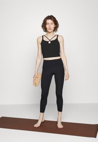 Top cropped preto com alças cruzadas e recorte, combinado com leggings pretas de cintura alta. Bloco de yoga de cortiça segurado em uma mão.