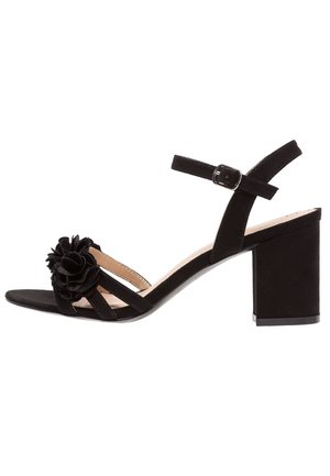 Schwarze Wildleder-High-Heelsandalen verfügen über ein florales Akzent, ein offenes Zehendesign, einen verstellbaren Knöchelriemen und einen klobigen Blockabsatz.