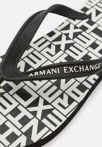 Svart gummislipper med teksturert såle, med et hvitt geometrisk mønster og "Armani Exchange"-logoen på stroppen.