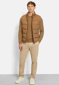 Braune, gepolsterte Weste über einem braunen Sweater, kombiniert mit beigen Hosen und hellen Sneakers. Glattes Material mit gestepptem Muster.