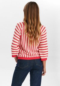 Sweter dzianinowy z wzorem w czerwono-kremowe paski, z rękawami o długości trzech czwartych oraz jednolitą, czerwoną ściągaczem na dole. Noszony z ciemnoniebieskimi dżinsami.