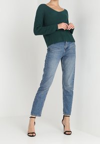 Pull côtelé vert foncé à col en V, associé à un jean taille haute bleu clair et des sandales à talons noires.