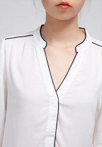 Blouse blanche avec un col noir bordé de piping et un décolleté en V. Le tissu semble lisse avec une texture légère. Aucun motif visible.