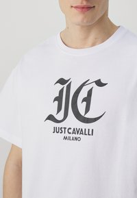 Bílý bavlněný tričko s velkým černým logem "JC" a textem "JUST CAVALLI MILANO" vytištěným na přední straně tučným písmem.