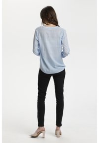 Frau mit langen Haaren, die sich abwendet, trägt ein hellblaues, gemustertes Langarmoberteil, schwarze Hose und beiges, hochhackiges Sandalen.