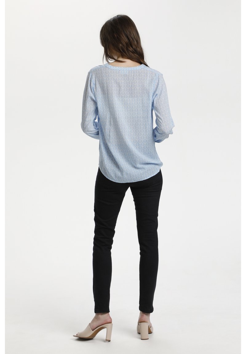 Frau mit langen Haaren, die sich abwendet, trägt ein hellblaues, gemustertes Langarmoberteil, schwarze Hose und beiges, hochhackiges Sandalen.