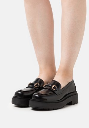 Svarta läderskor med plattform och gyllene spännen, barfota på en vit bakgrund.