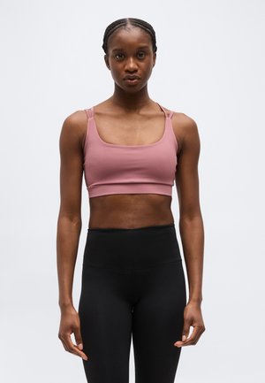 Športovo vyzerajúca žena s vrkočmi, oblečená v purpurovom športovom podprsenke a čiernych legínach s vysokým pásom, stojaca pred jednoduchým pozadím.