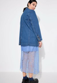 Chaqueta de mezclilla en azul medio con un corte relajado, superpuesta sobre un vestido de encaje azul claro. El encaje tiene un patrón floral y un dobladillo en forma de escallola.