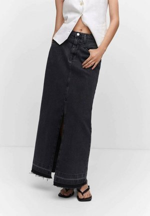 Woman wearing a high-waisted black denim maxi skirt with front slit and frayed hem, paired with black sandals and a white sleeveless top.