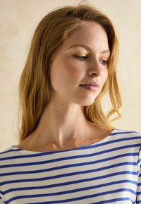 Ribaga ülaosa laia ja merevaigukollase kaelusega valges ja sinises. Pehme kangas, keha lähedal disain, rõhutades lõdva siluetti. Loomulik nahatooniga taust.