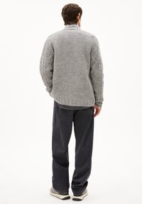 Grauer Strickpullover mit hohem Kragen und strukturiertem Zopfmuster, kombiniert mit dunklen, weit geschnittenen Hosen und lässigen Schuhen.