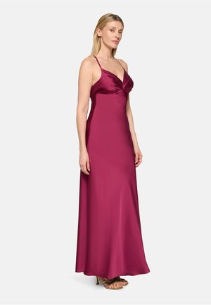 Femme debout de profil portant une robe de soirée en satin rose profond sans manches, avec des bretelles fines, et des talons ouverts dorés, sur un fond blanc.