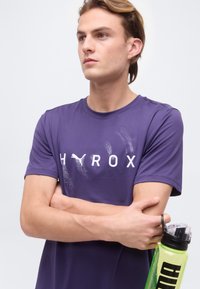 Νέος άνδρας με μωβ αθλητική μπλούζα Puma και σταυρωμένα χέρια κρατά πράσινο παγούρι Puma, κοιτάζοντας προς το πλάι μπροστά σε απλό φόντο.