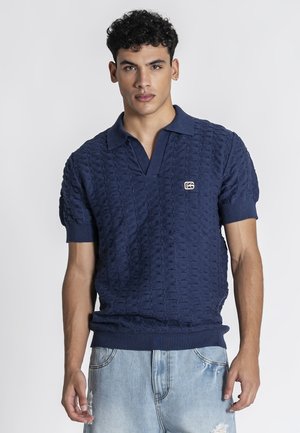 Giovane uomo con capelli neri ricci che indossa un maglione polo a maniche corte in blu navy con trama e pantaloncini in denim chiaro consumati.