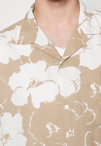 Camicia con maniche corte in tessuto leggero, con motivo floreale beige e bianco, dotata di colletto a V e una tasca sul petto.