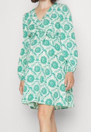 Kvinde iført en knælængde, langærmet kjole med grønne blomsterprint og bundet sløjfe foran i taljen.