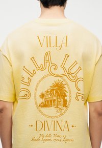 Άτομο που φοράει ανοιχτόκίτρινη μπλούζα με την επιγραφή «Villa Della Luce Divina» και γραφικό ενός αρχοντικού με φοίνικα στην πλάτη.