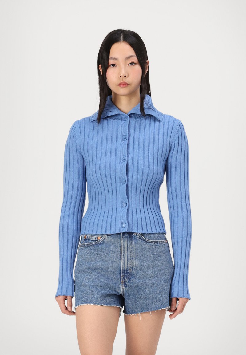 Cardigan bleu côtelé avec col, fermeture par boutons et manches longues. Porté sur un short en denim, avec une coupe courte et un tissu texturé.