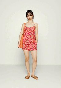 Macacão com estampa floral vermelha e alças finas, apresentando calções folgados. Acompanhado por uma bolsa tote laranja e sandálias tipo slide.