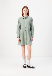 Abito button-up verde chiaro con maniche lunghe, un corpino aderente e una gonna svasata. Indossato con scarpe basse nere e calzini bianchi alla caviglia.