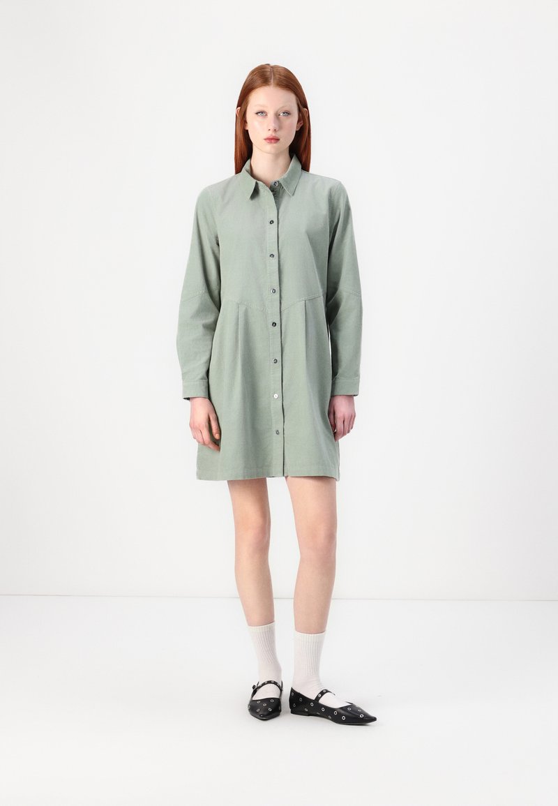 Abito button-up verde chiaro con maniche lunghe, un corpino aderente e una gonna svasata. Indossato con scarpe basse nere e calzini bianchi alla caviglia.